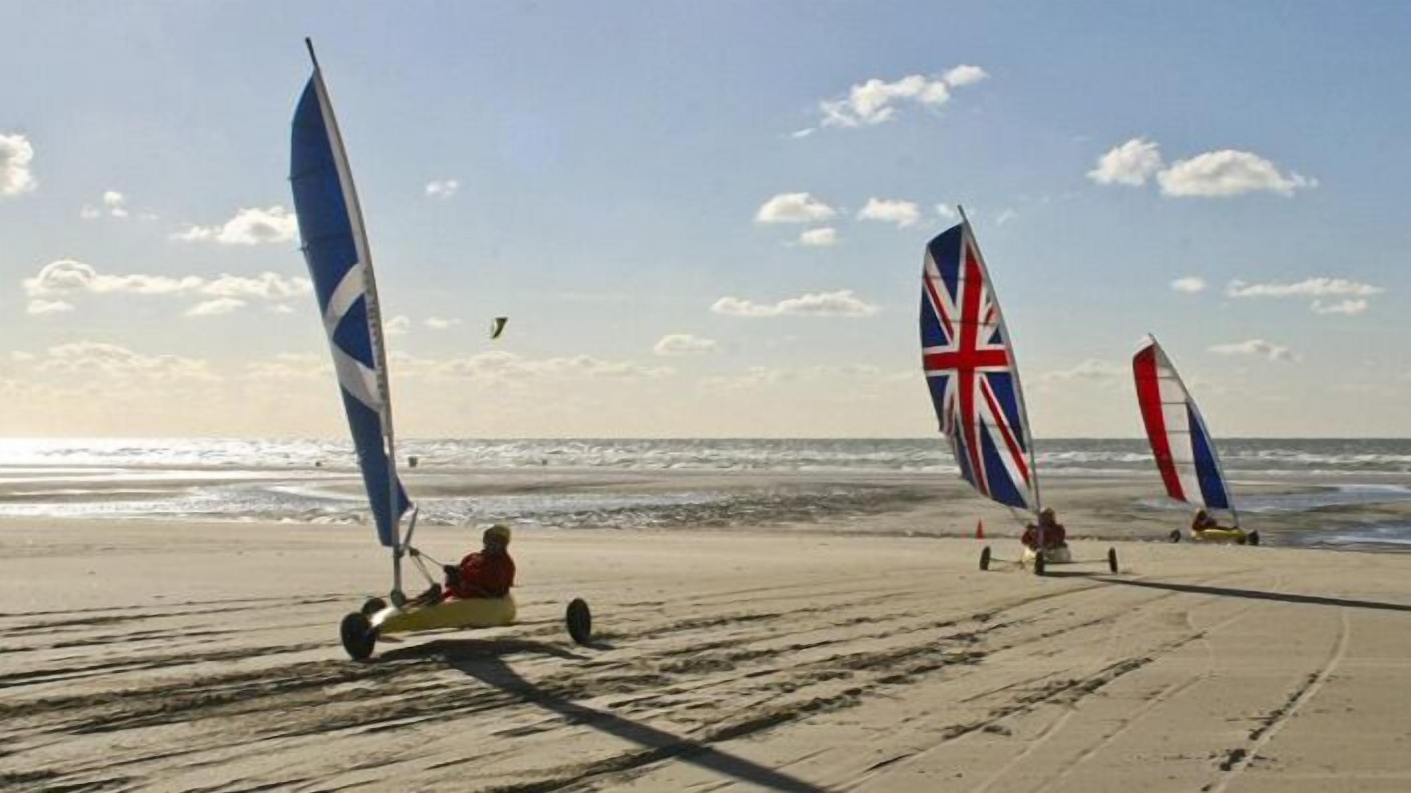 A beach with three sailboards, one with a blue sail and another with a Union Jack flag.
