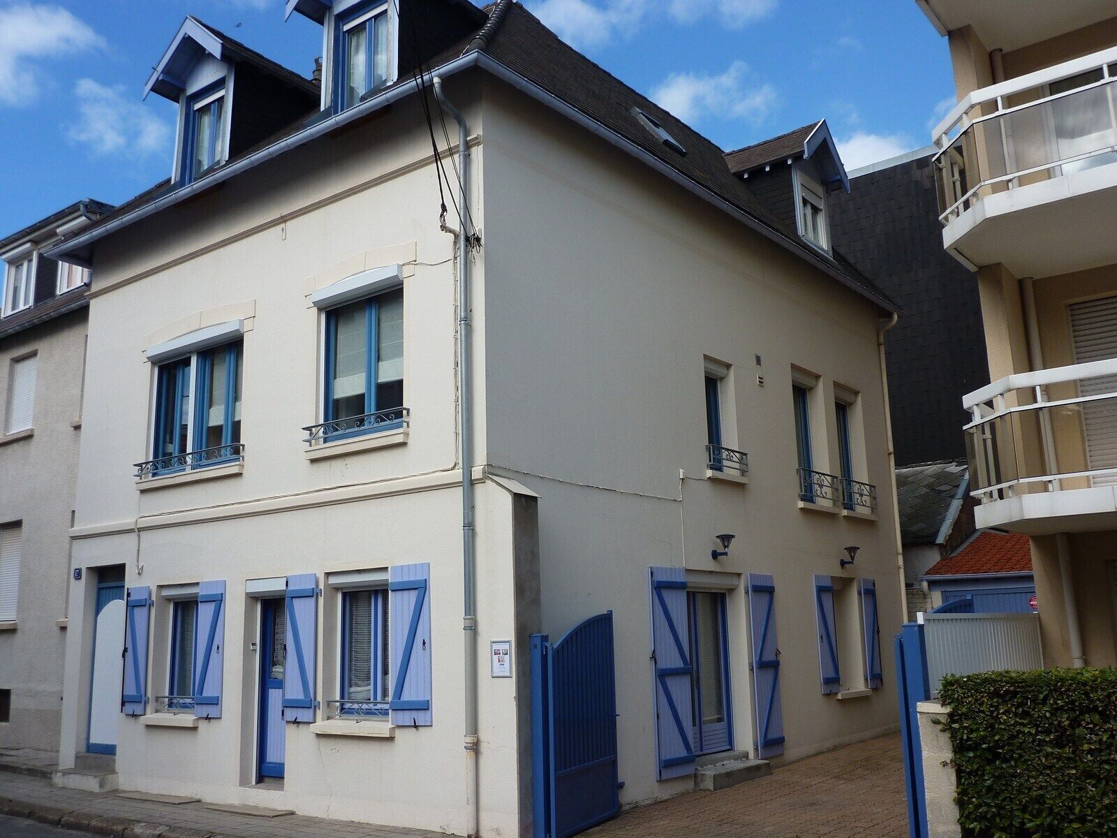 A two-story building with blue shutters, a balcony on the second floor, and a gated entrance.