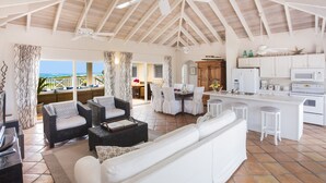 A spacious living area with a white sofa, wicker chairs, and a dining table. A kitchen with white cabinets and a large island is visible.
