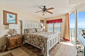 A bedroom with a bed, a nightstand, a dresser, a ceiling fan, a window with blinds, and a view of the ocean.