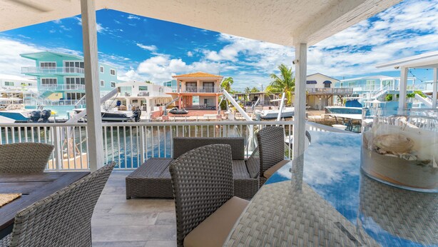 A covered outdoor seating area with wicker furniture, a glass table, and a view of a marina with boats and buildings. - Sandbars/Fishing/Boat Rental Dreamy Keys Gateway Home On The Water! (Tavernier)