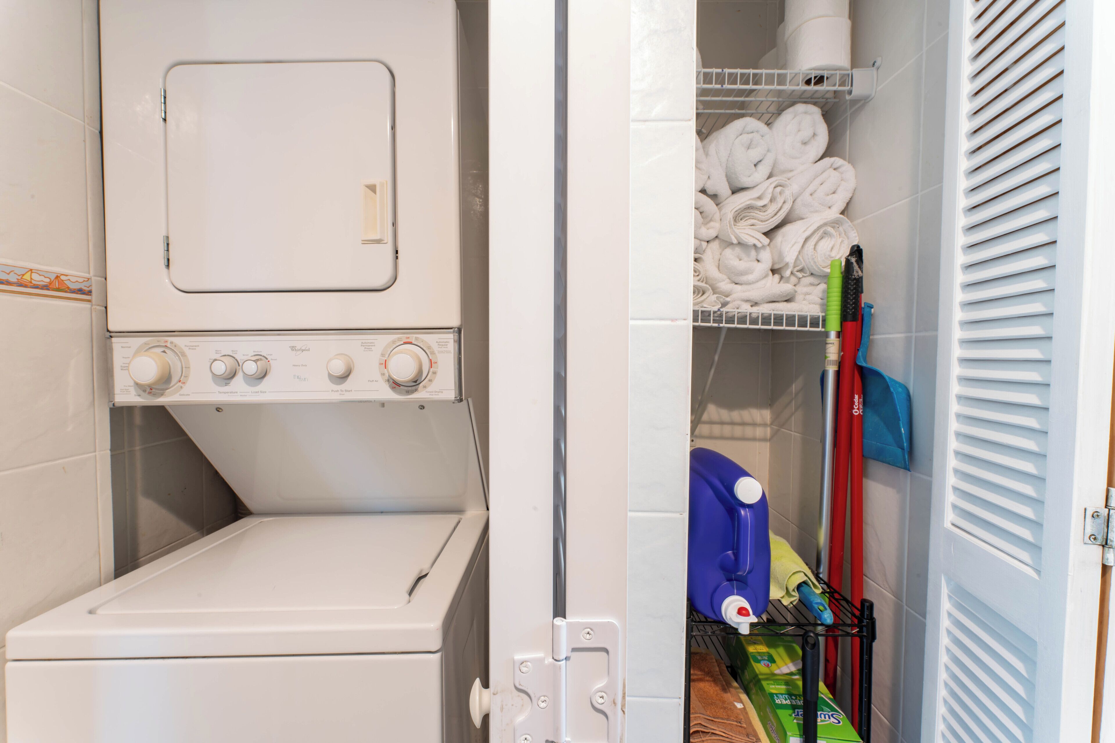 A laundry room with a front-loading washing machine, a drying rack with folded towels, and a shelf with cleaning supplies.