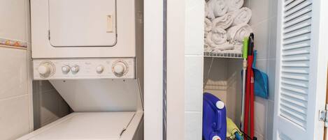 A laundry room with a front-loading washing machine, a drying rack with folded towels, and a shelf with cleaning supplies.