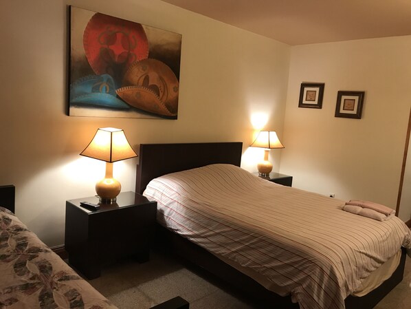 Una habitación de hotel con una cama, dos mesitas de noche con lámparas, un cuadro en la pared y dos fotografías enmarcadas.