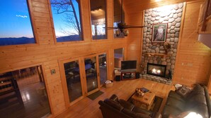 A spacious living area with a fireplace, wooden walls, and large windows offering a view of the outdoors. - HAWKS RIDGE HIDEAWAY- Luxury Cabin & Great Get-Away! Amaazing view! (Bryson City)