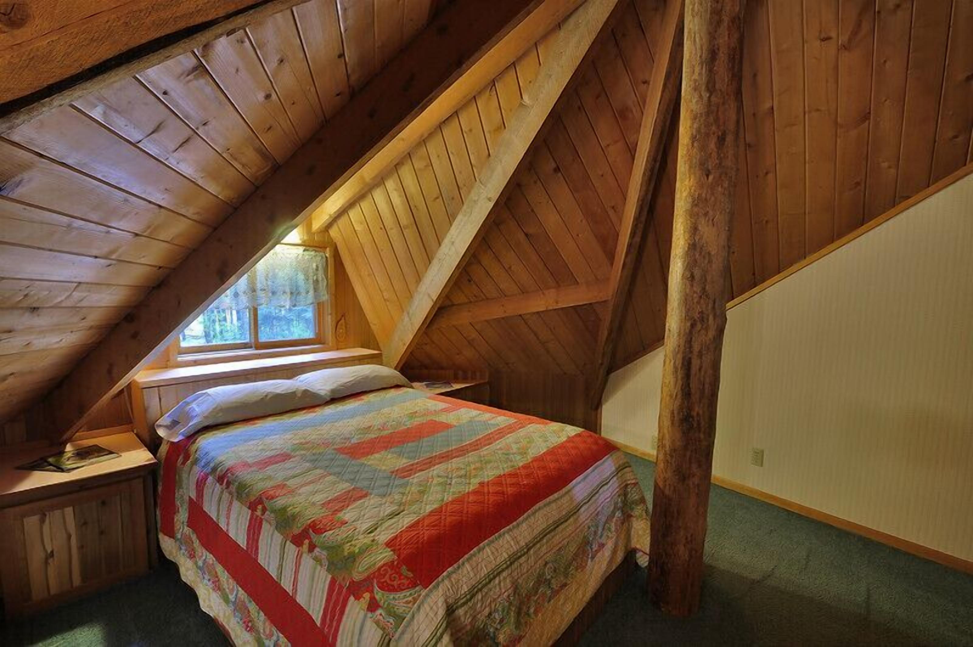 Um quarto de sótão acolhedor com teto de madeira inclinado, uma cama de solteiro com um edredom colorido e uma pequena mesa de cabeceira.