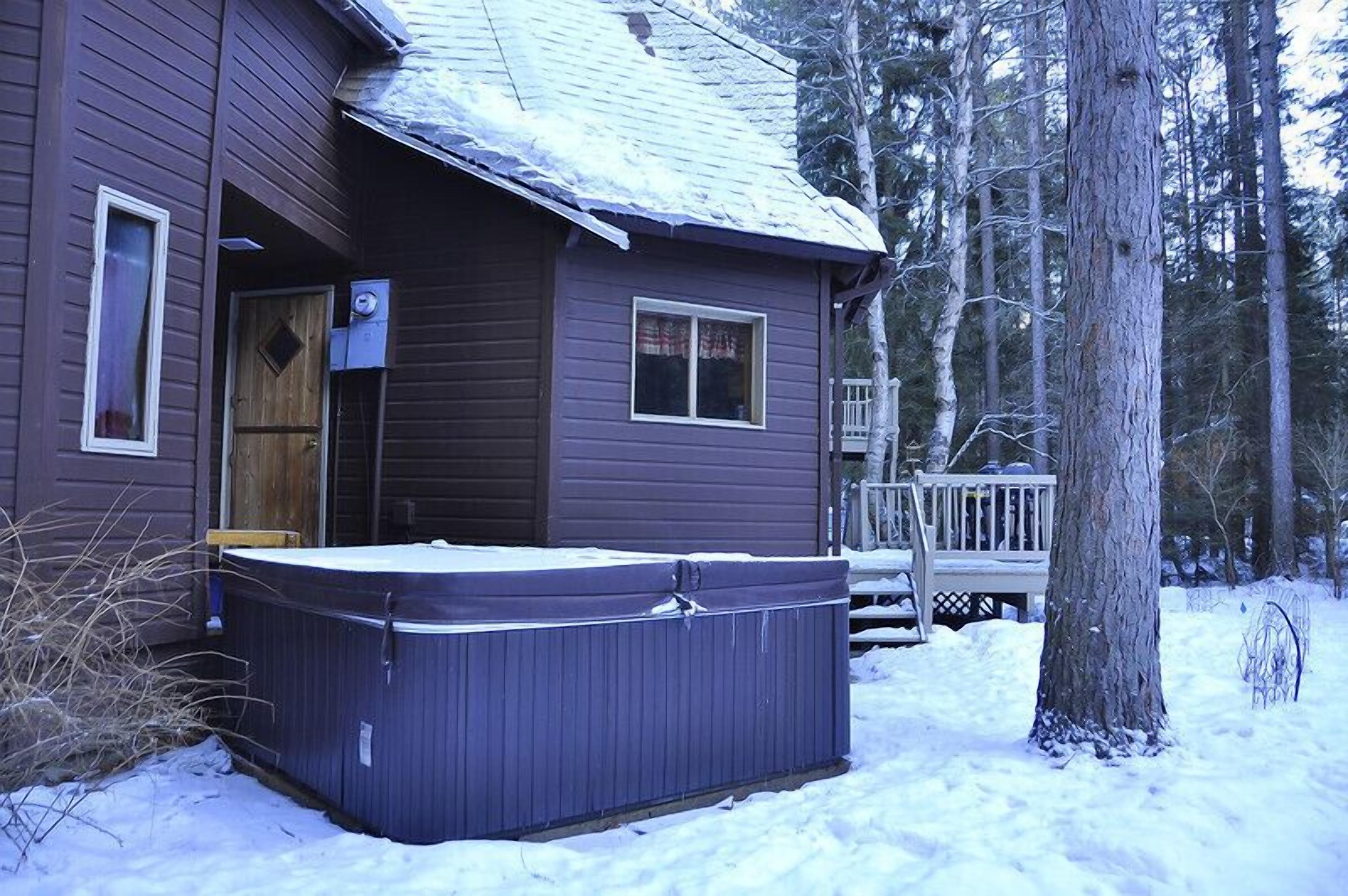 A wooden cabin with a hot tub in front, surrounded by snow and trees.