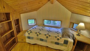 Une chambre mansardée chaleureuse, avec un plafond en pente en bois, un lit recouvert d’une courtepointe, deux petites fenêtres et une table de chevet avec une lampe.