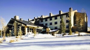 A large, multi-story building with a modern architectural style, featuring a mix of stone and glass facades, surrounded by snow-covered ground and sparse trees. - Brian head Studio in 3.5 star Cedar Breaks Lodge - Sleeps 4 (Brian Head)