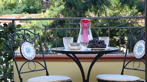 Una mesa redonda con una botella, un cuenco y un plato de uvas en un balcón con sillas de metal.