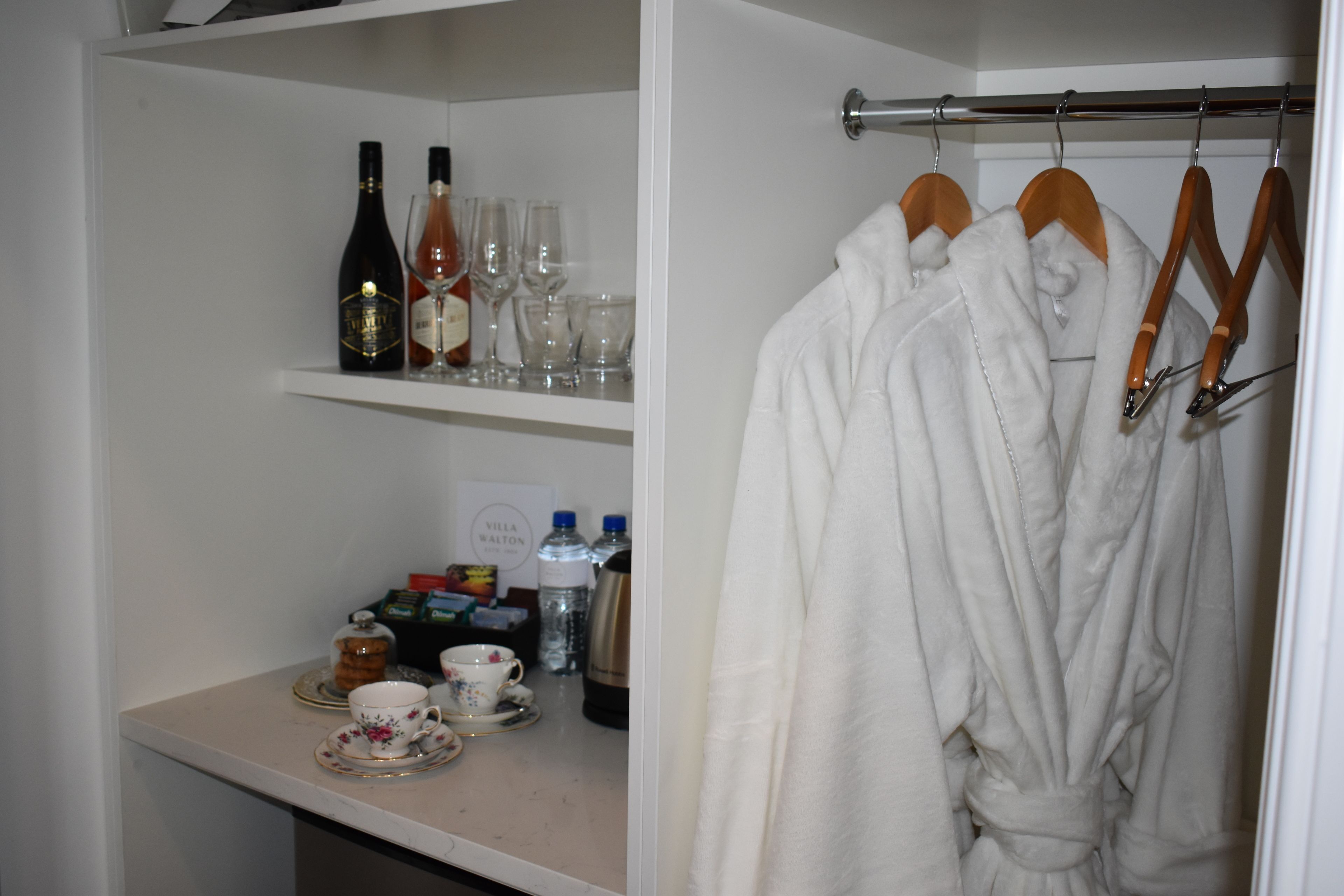 A hotel room with a minibar, a robe hanging on a rack, and a shelf with bottles and glasses.