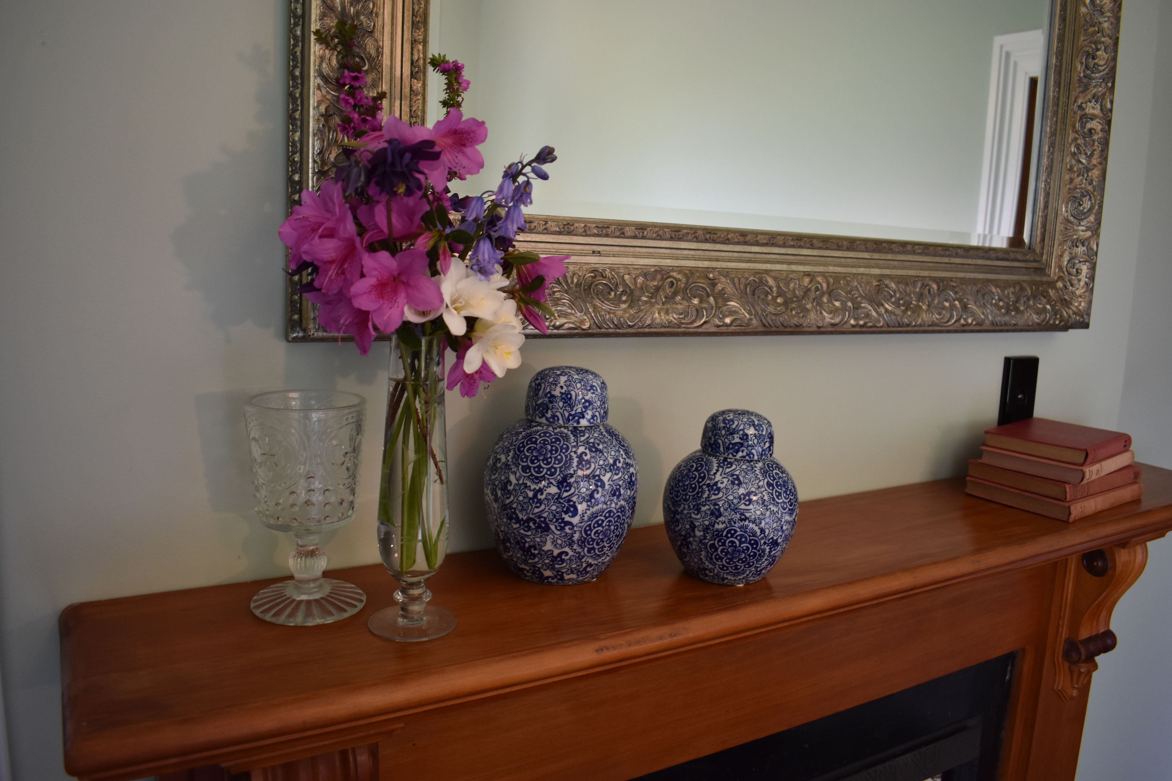 A wooden mantelpiece with a vase of flowers, a glass, and two blue patterned vases.