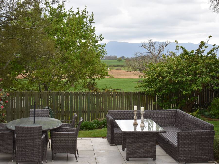 Un patio con mobiliario de mimbre y una mesa de vidrio, rodeado por una cerca de madera y follaje, con vistas a un campo y montañas distantes.
