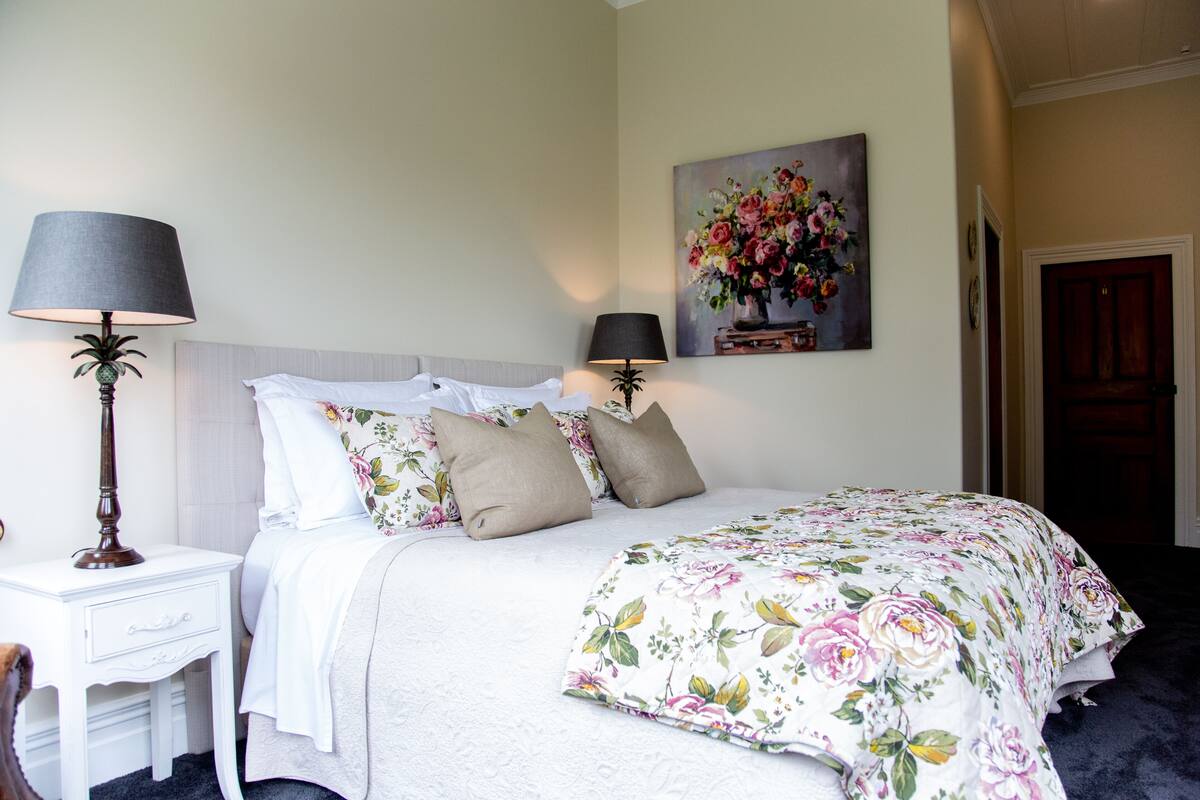 A neatly made bed with floral bedding, two bedside lamps, a small bedside table, and a floral painting on the wall.