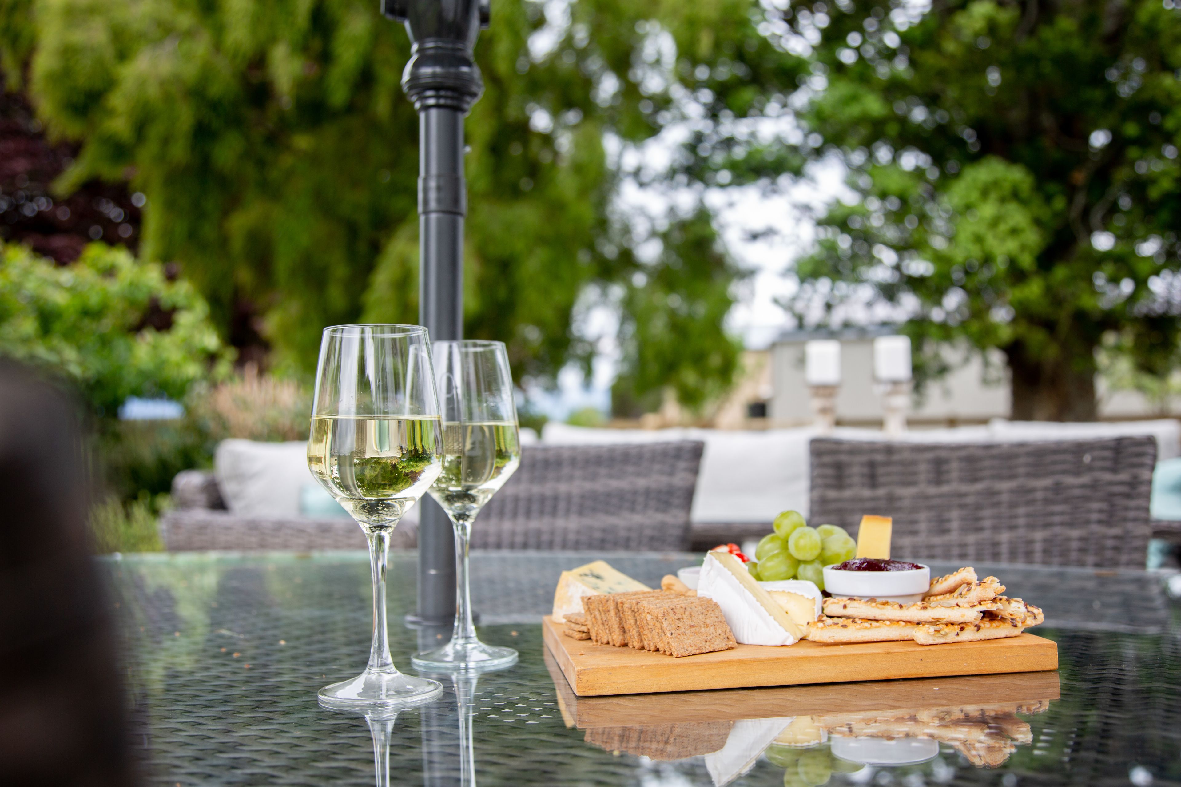 Una mesa de vidrio con dos copas de vino, una tabla de madera con queso, galletas, uvas y un recipiente pequeño de mermelada.