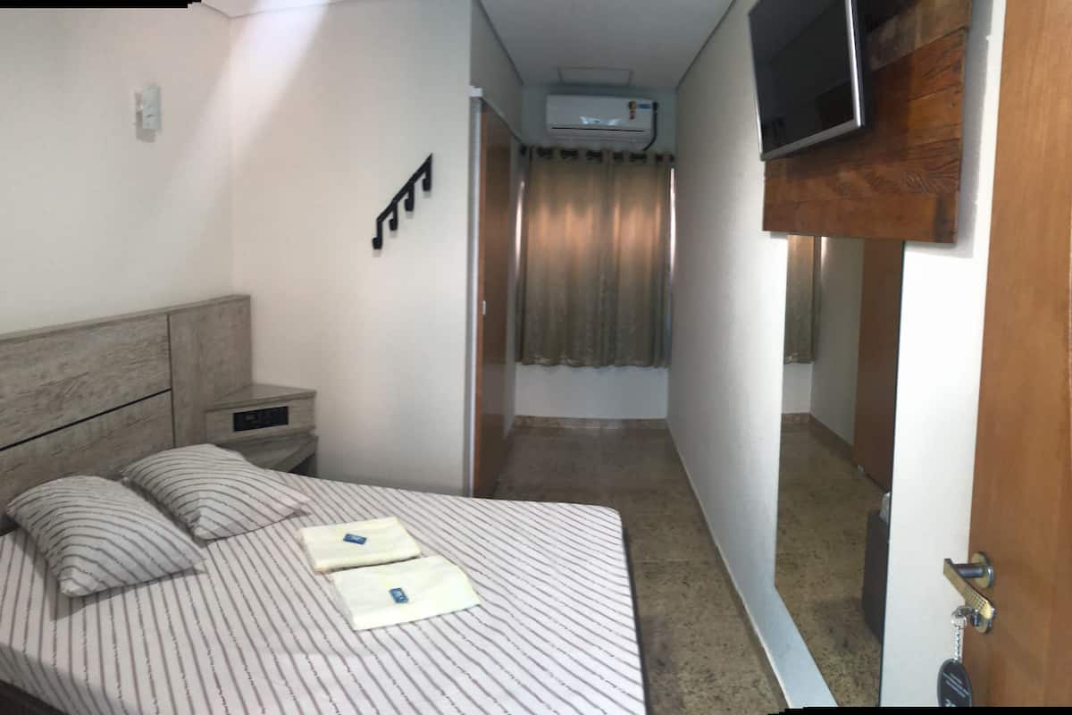 A hotel room with a bed, a nightstand, a TV mounted on the wall, and an air conditioning unit.