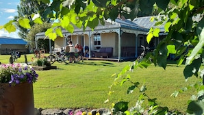 A house with a covered porch, a lawn, and a garden with flowers. - Peter's Farm Lodge (Waipiata)