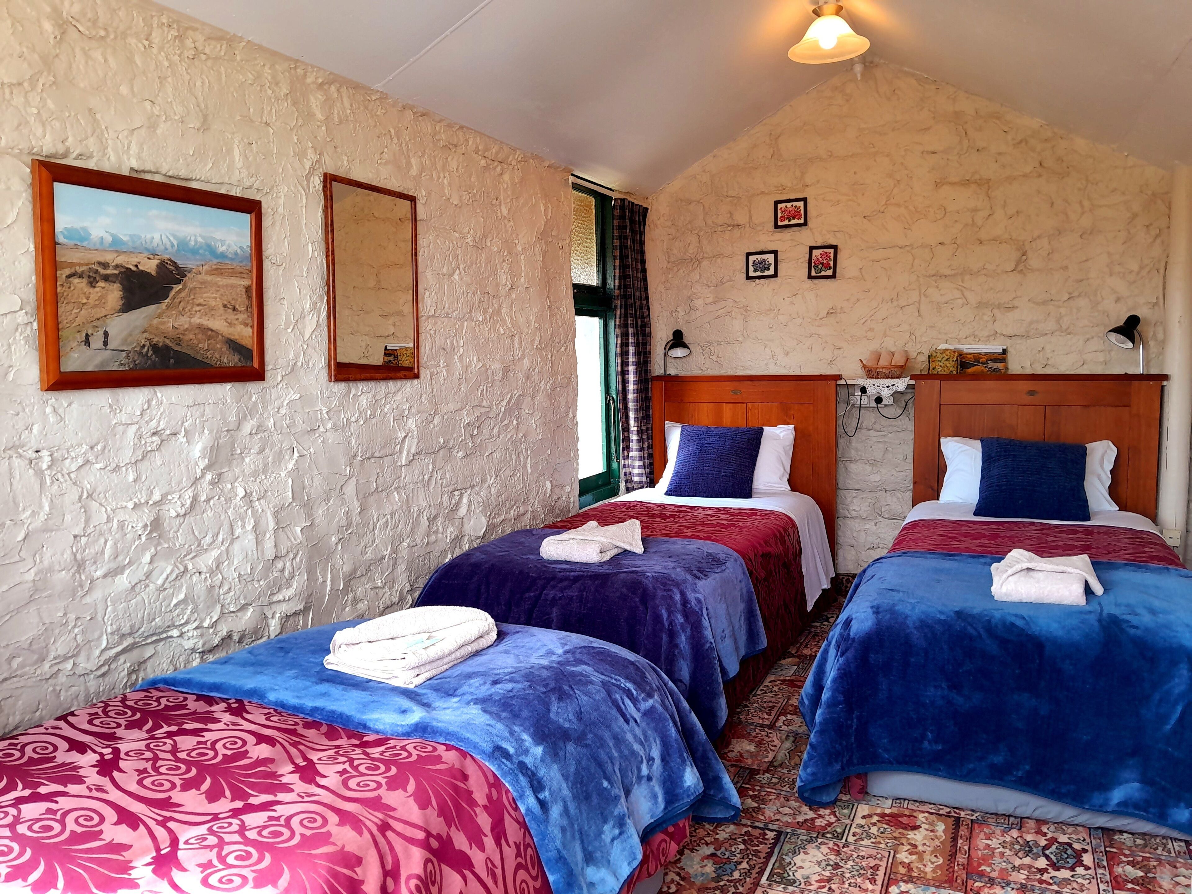 A room with two beds, each with a blue and red bedspread, a wooden headboard, and a painting of a mountainous landscape on the wall.