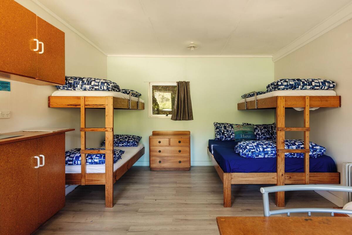 A dormitory room with bunk beds, a wooden dresser, a small table, and a window with curtains.