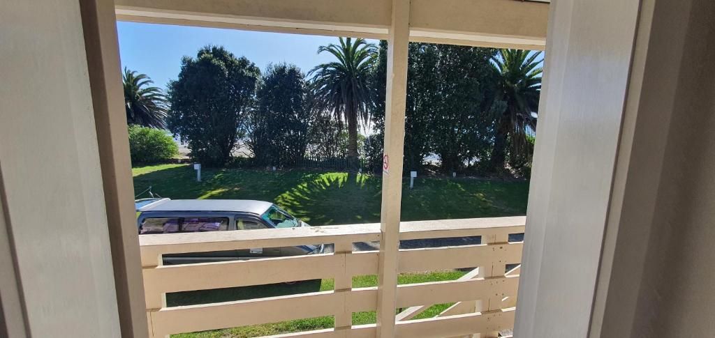A view through a window with a wooden railing, a white car parked outside, and palm trees in the background.