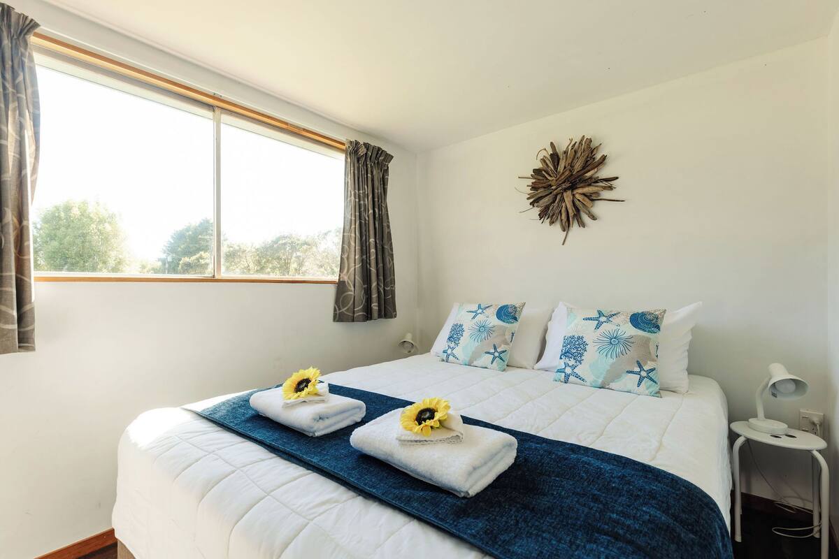 A neatly made bed with a blue blanket and two towels with sunflowers, a decorative wall piece, and a window with curtains.