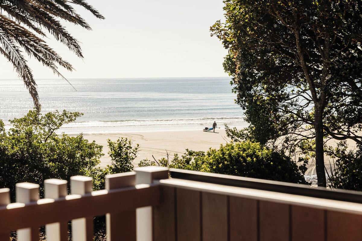 A beach view from a balcony with a wooden railing, a person walking on the beach, and a palm tree in the foreground.