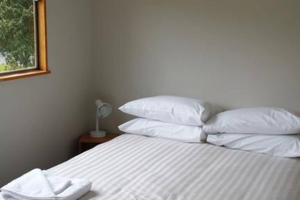 A neatly made bed with white linens, a white lamp on a bedside table, and a window with a view of trees.