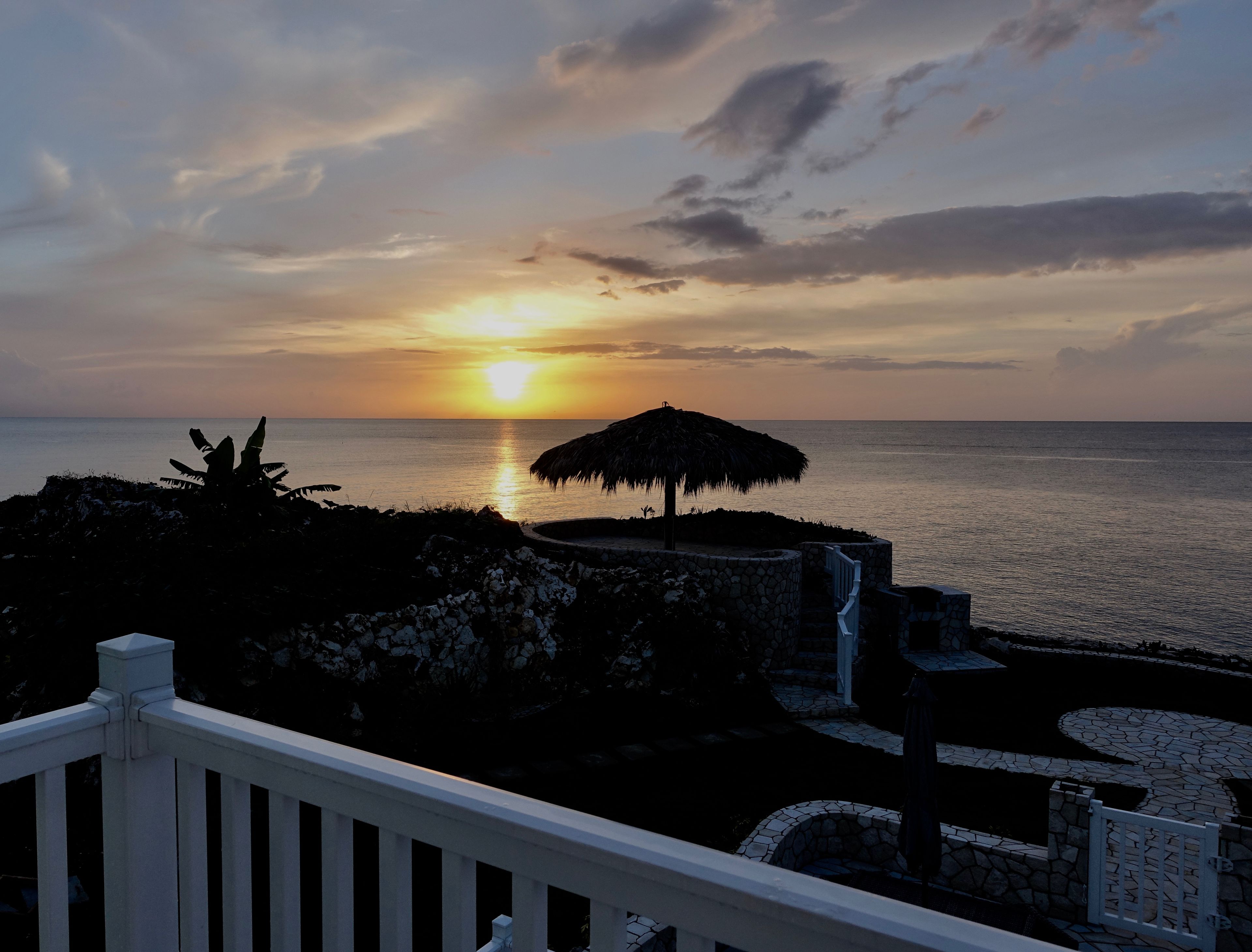 Une vue sur le coucher de soleil depuis un balcon donnant sur la mer, avec un parasol en chaume et des marches de pierre menant à l’eau.