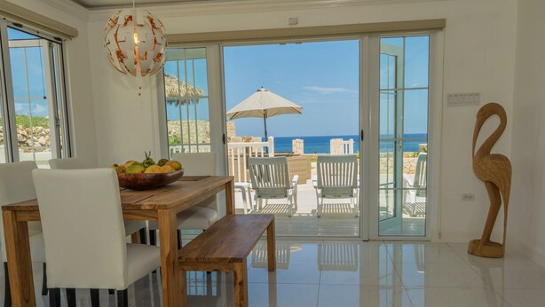 A dining area with a wooden table and chairs, a fruit bowl, and a view of the ocean through large glass doors. - Stunning Ocean View (Negril)