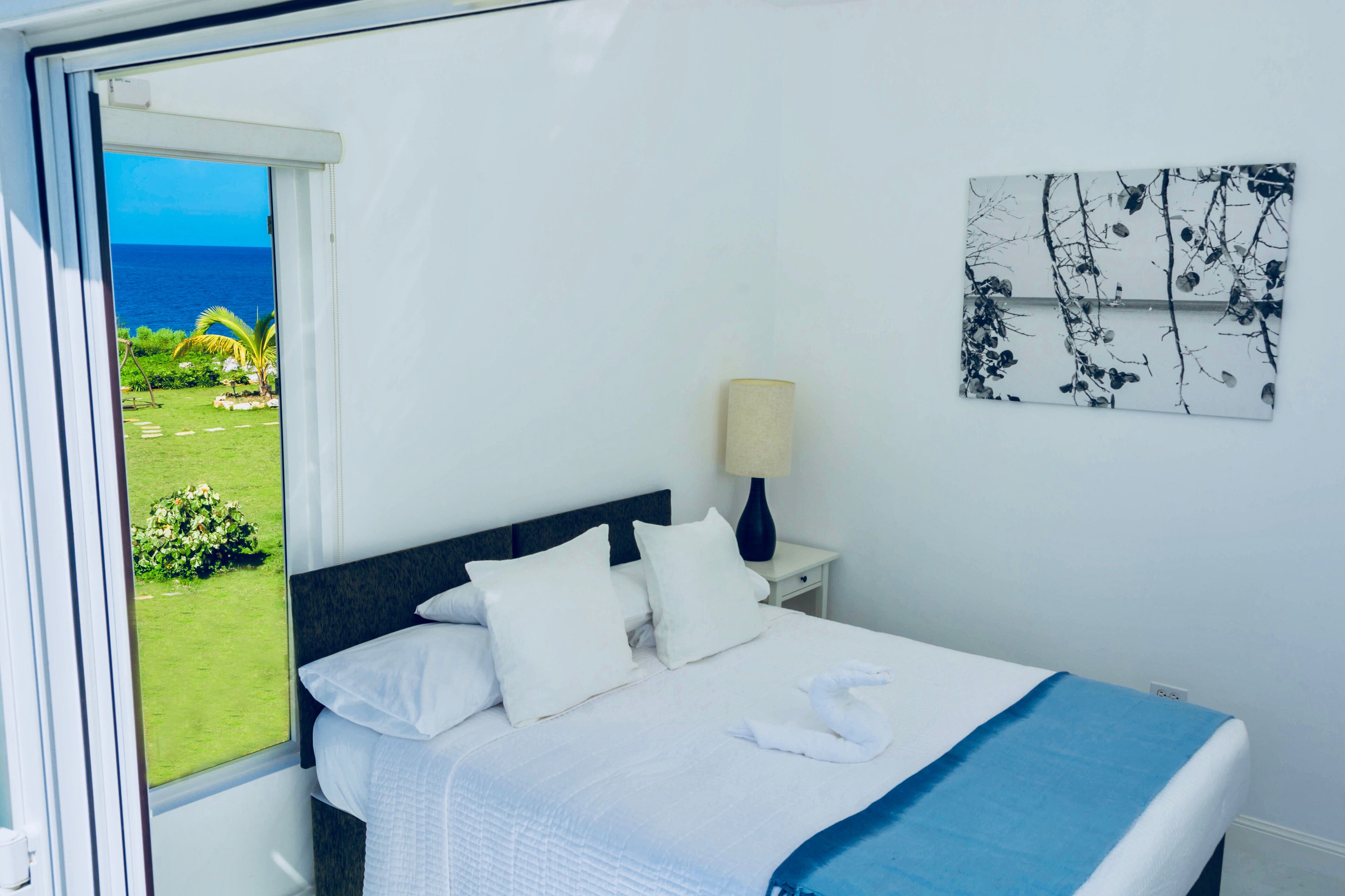 Una camera d'albergo con un letto, un comodino, una lampada, una finestra vista mare e vegetazione, e un'opera d'arte appesa al muro.