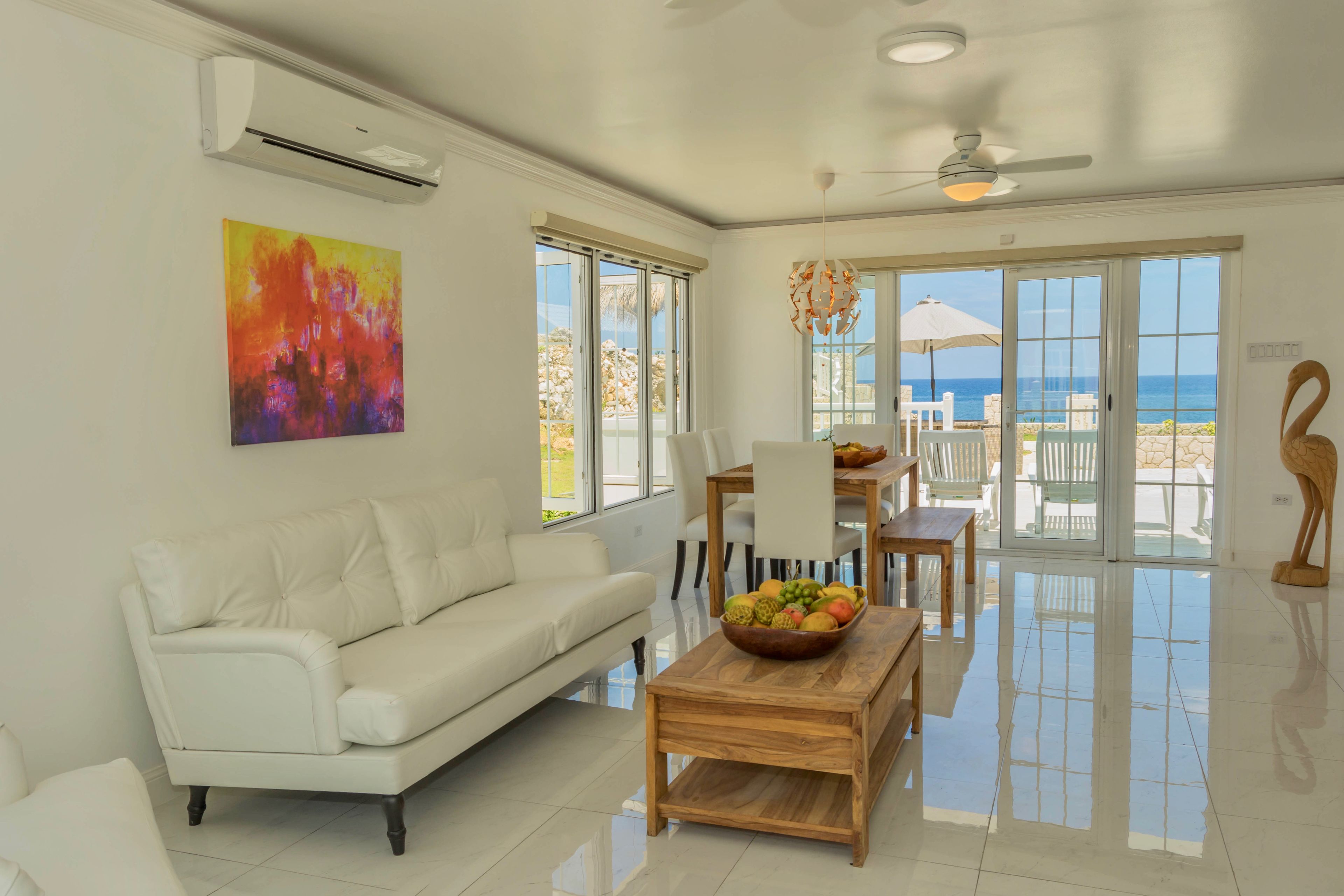 A modern living room with a white sofa, a wooden coffee table with fruit, and a view of the ocean through large sliding doors.