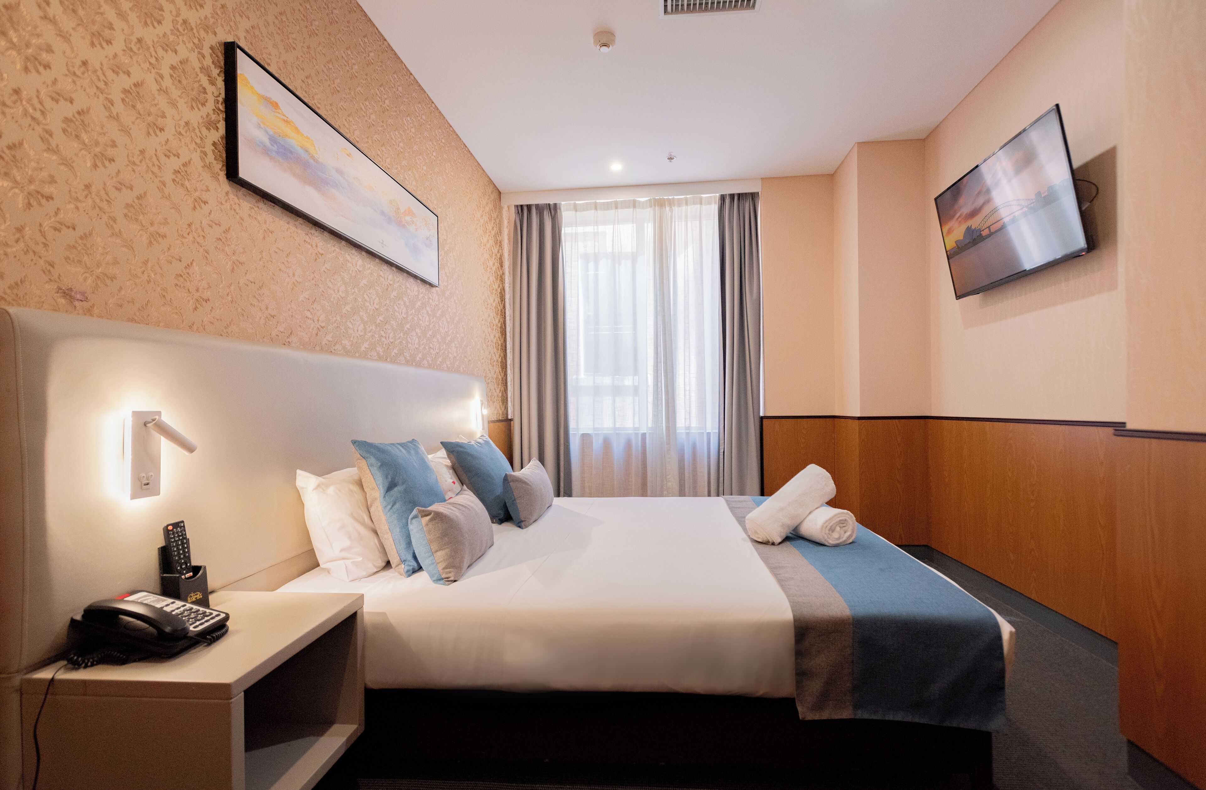 Une chambre d’hôtel avec un lit bien fait, une table de chevet avec un téléphone et une télécommande, un téléviseur fixé au mur et une fenêtre avec des rideaux.