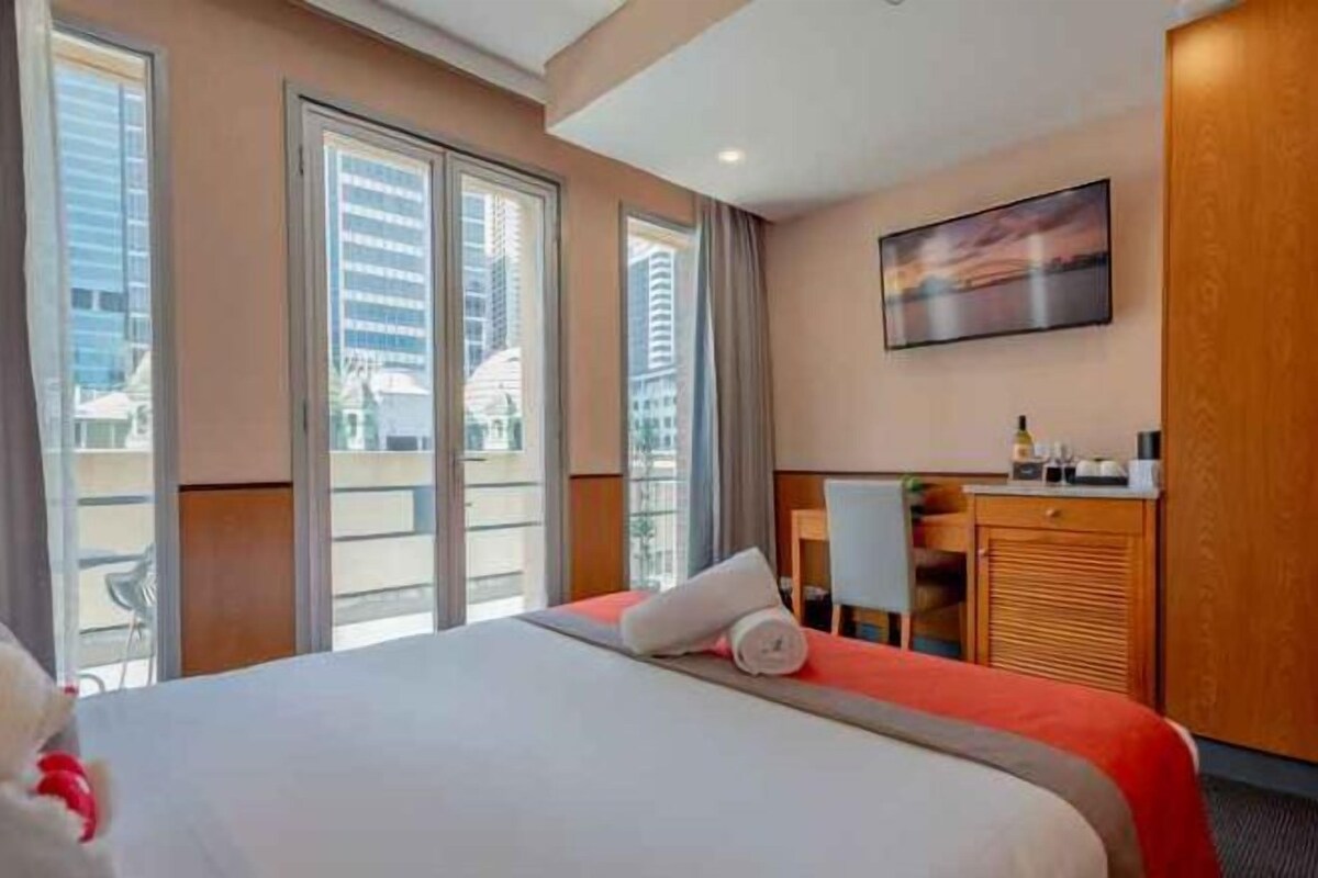 A hotel room with a large bed, a desk, and a view of the city through large windows.