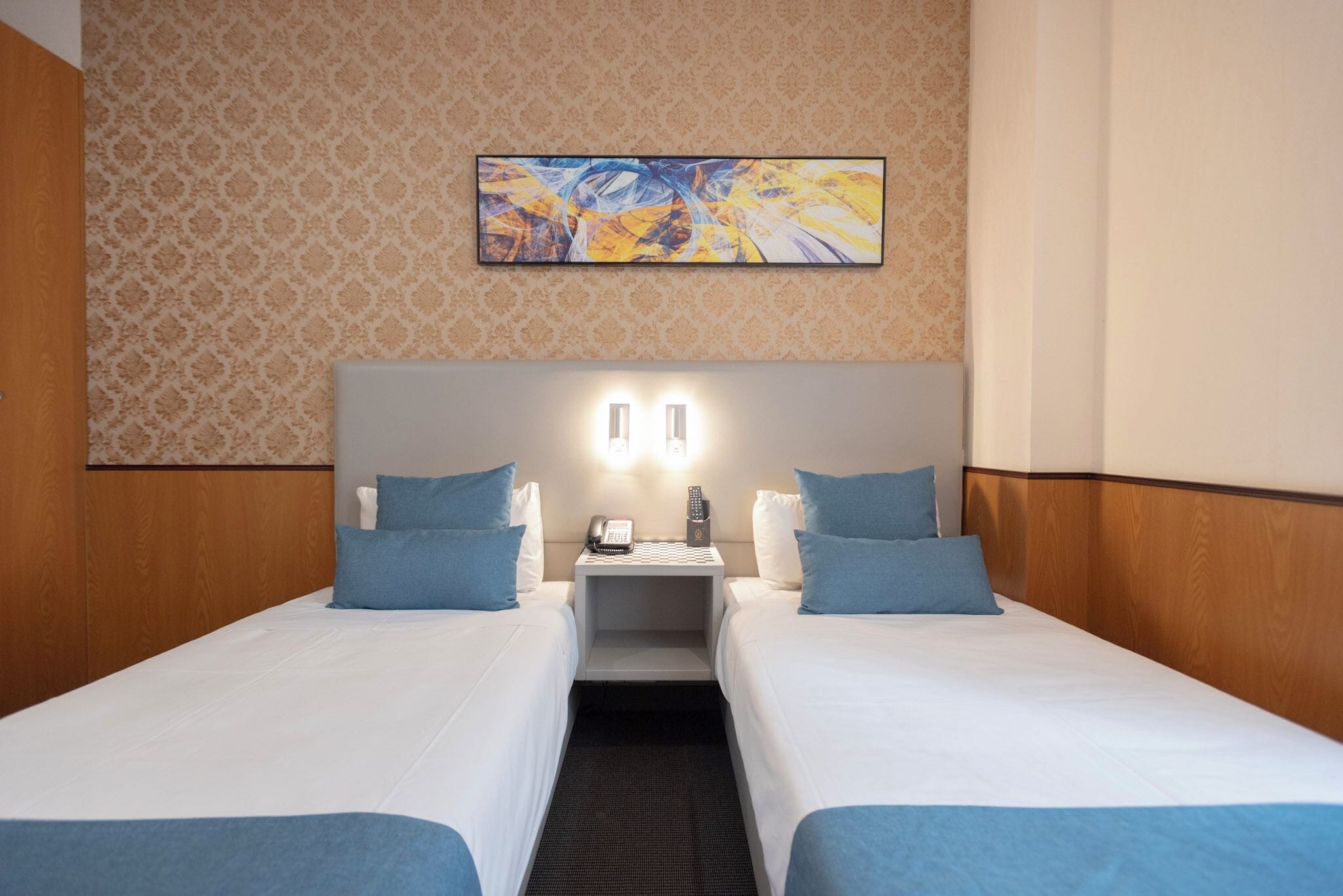 Habitación de hotel con dos camas, una mesita de noche con teléfono y lámpara, y un cuadro abstracto colgado en la pared.