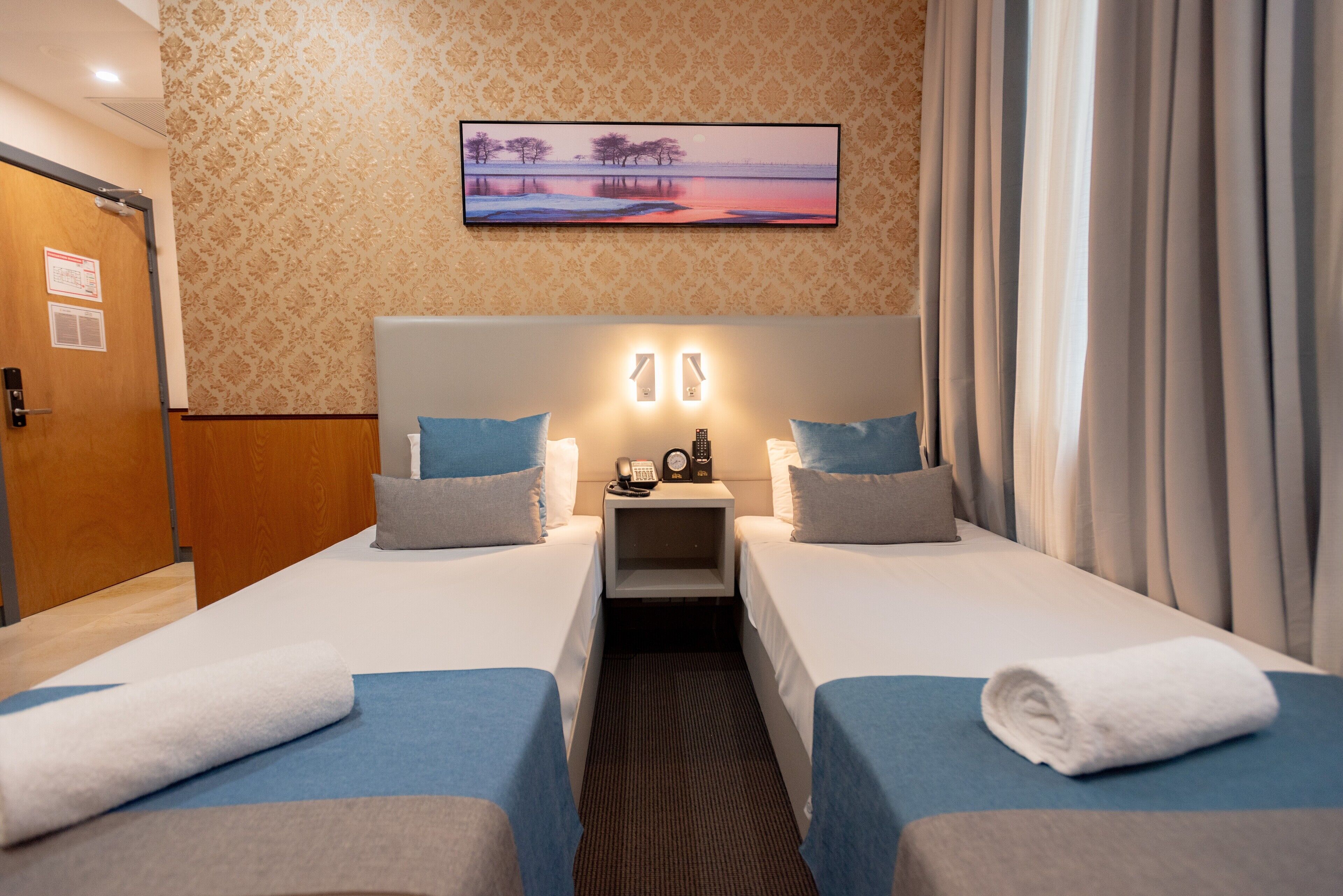 A hotel room with two beds, a nightstand with a phone and a clock, and a painting on the wall.