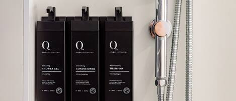 Three black bottles of Q oxygen collection products: shower gel, conditioner, and shampoo, mounted on a wall next to a shower.