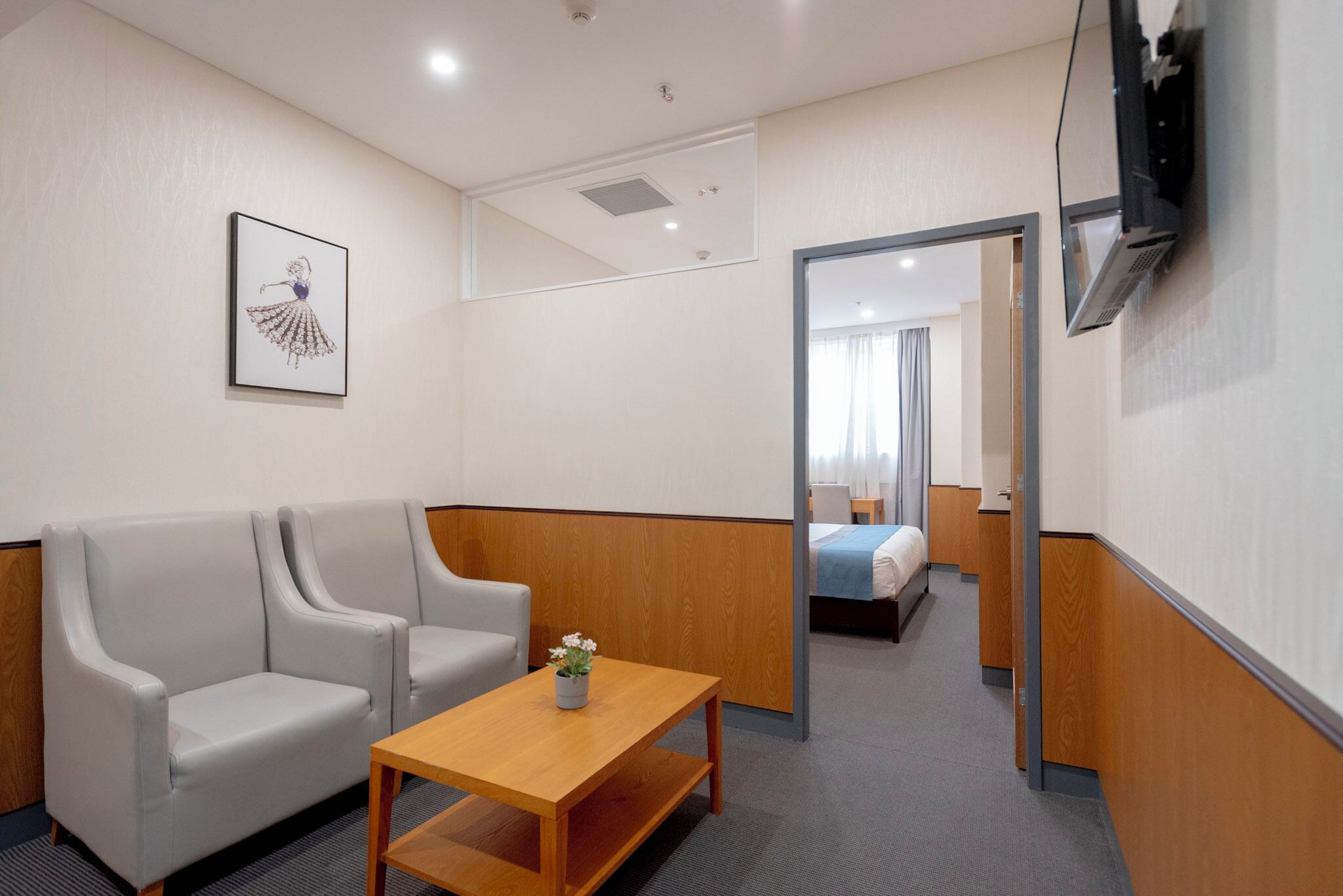 A hotel room with a sofa, a wooden table, a TV mounted on the wall, and a bed in the background.