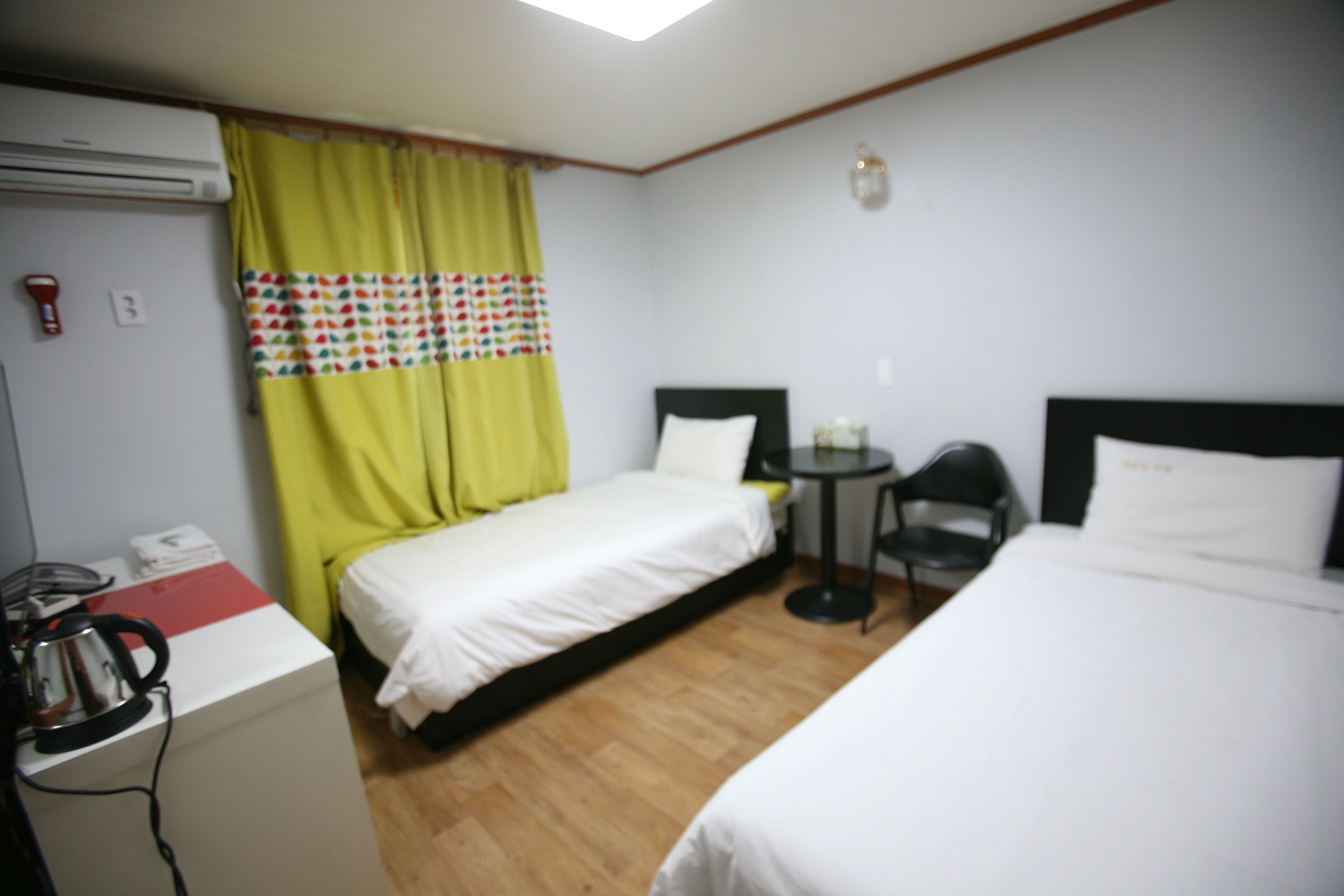 Habitación de hotel con dos camas individuales, una mesita, una silla y una zona de kitchenette con hervidor.