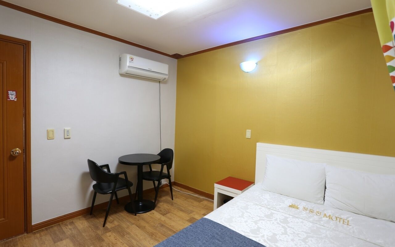 Una habitación con cama, una mesita pequeña con dos sillas, un equipo de aire acondicionado y una lámpara fijada a la pared.