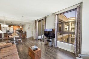 Una sala de estar moderna con televisor grande, piso de madera y un comedor con cocina al fondo.