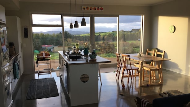 A modern kitchen with a large island, dining area with wooden chairs, and a view of the countryside through large windows. - Land escape in Bream Bay (Waipu)