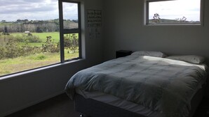A bedroom with a large bed, two windows, and a view of a green landscape. - Land escape in Bream Bay (Waipu)