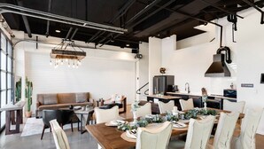 Un espace de salle à manger moderne, avec une table en bois dressée pour six personnes, entourée de chaises rembourrées. Un lustre est suspendu au plafond, et on aperçoit un coin cuisine à l’arrière-plan.
