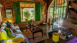 A cozy room with a brick wall, green shutters, a wooden sofa, a coffee table, and a large window with a view of greenery.
