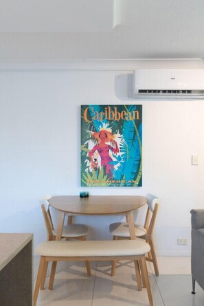 Un coin repas avec une table ronde en bois et quatre chaises, une affiche des Caraïbes accrochée au mur et un canapé gris.