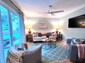 A living room with a sofa, armchairs, a coffee table, a ceiling fan, and a TV mounted on the wall.