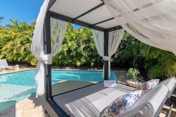 Ein privater Poolbereich mit Liegebett, Lounge-Stühlen und üppiger Bepflanzung im Hintergrund.