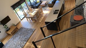 A modern kitchen with a built-in oven, a dining area with a wooden table and chairs, a large window overlooking a backyard, and a staircase leading to an upper level. - Modern Beach-House Gem (Dodges Ferry)