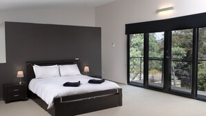 A modern bedroom with a large bed, two bedside tables with lamps, and a view of greenery through large windows. - Modern Beach-House Gem (Dodges Ferry)