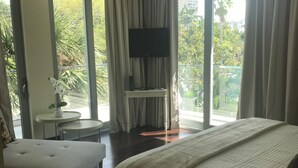 Hotelkamer met een groot bed, een zithoek met een tafeltje, een wandgemonteerde flatscreen televisie en grote ramen met uitzicht op groen en gebouwen.